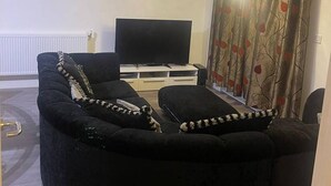 Un saló amb un sofà negre, una televisió de pantalla plana, un rellotge a la paret i una finestra amb cortines.