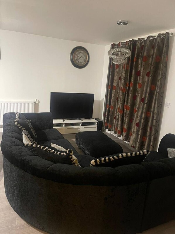 Un salón con un sofá negro curvo, un televisor de pantalla plana, un mueble blanco y una cortina estampada.