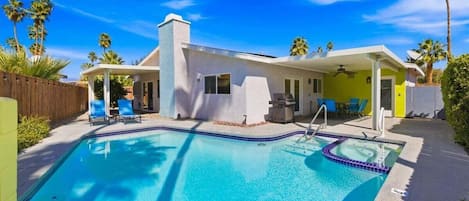 A pool area with a house, a covered patio, and palm trees.