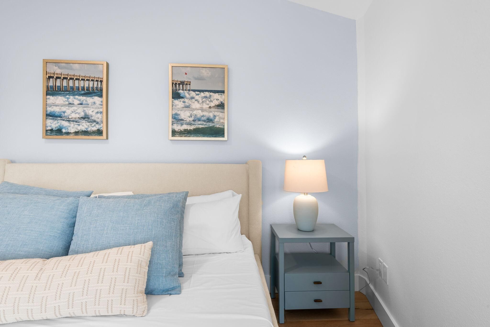 Un dormitorio con cama, almohadas, una mesita de noche con lámpara y dos cuadros en la pared.