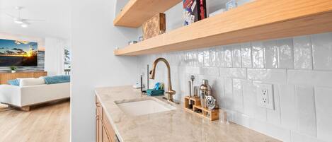 A modern kitchen with a marble countertop, wooden shelves, and a view of the living area.