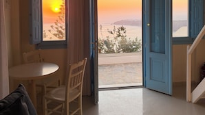 Habitació amb porta oberta que dona a un balcó, taula i cadires, i vista al capvespre.