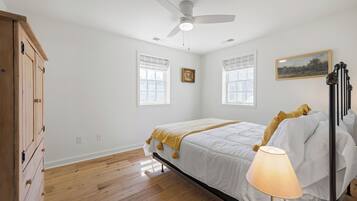 A bedroom with a bed, a ceiling fan, two windows with curtains, a bedside lamp, and a wooden wardrobe.