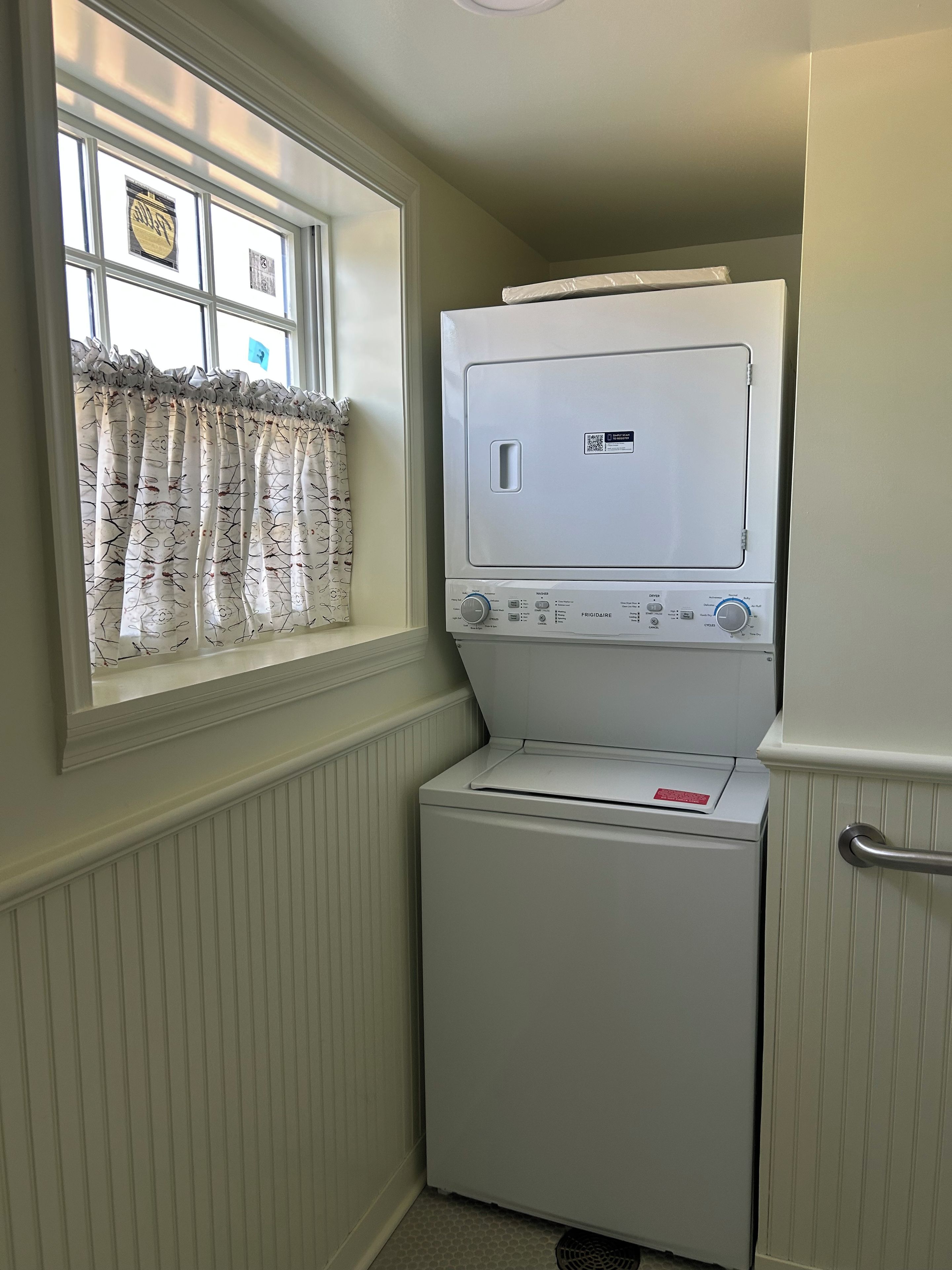 A compact laundry room with a front-loading washer and dryer, a window with a curtain, and a door with a handle.