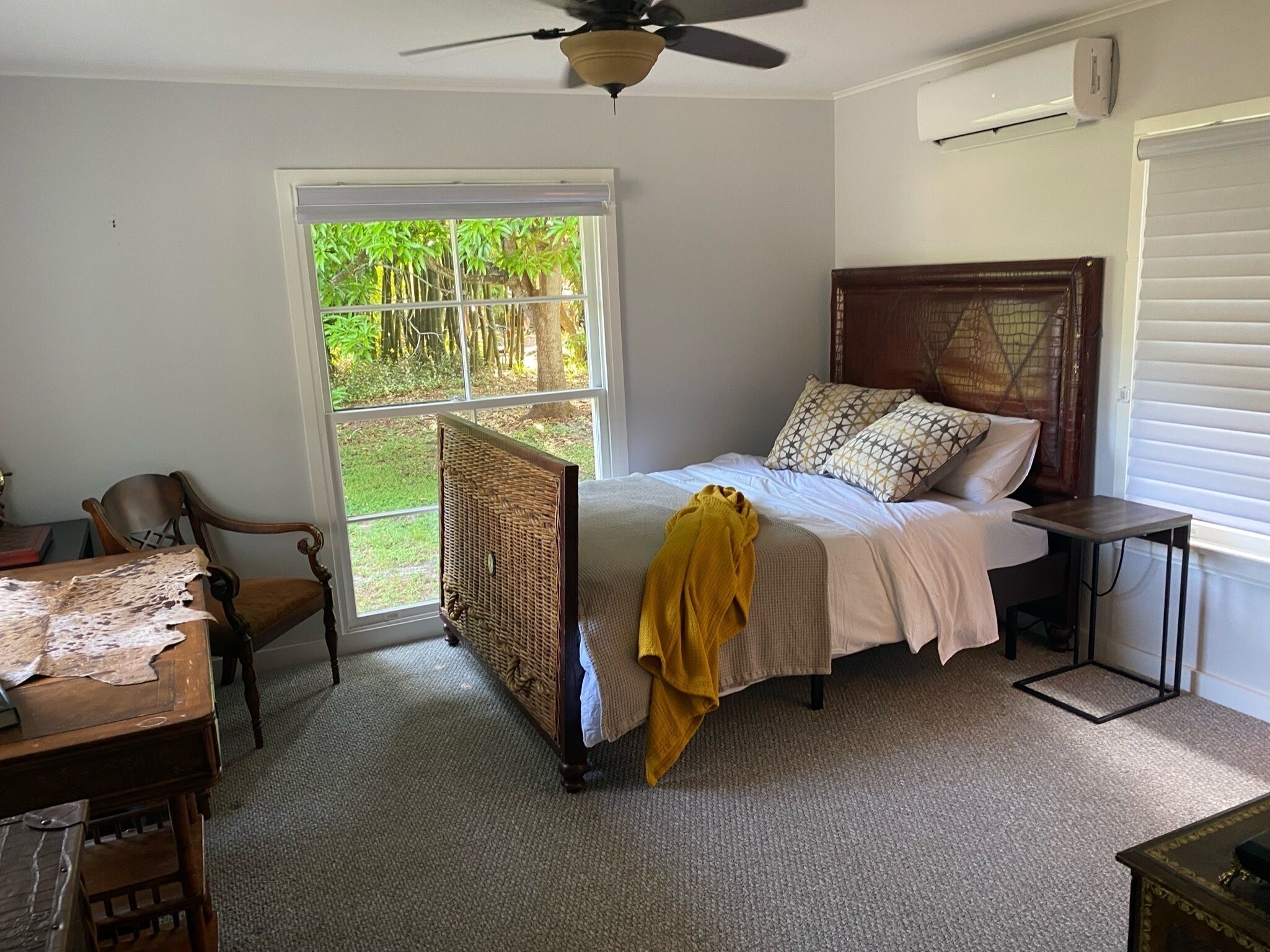 A bedroom with a bed, a chair, a small table, a window with a view of greenery, and a ceiling fan.