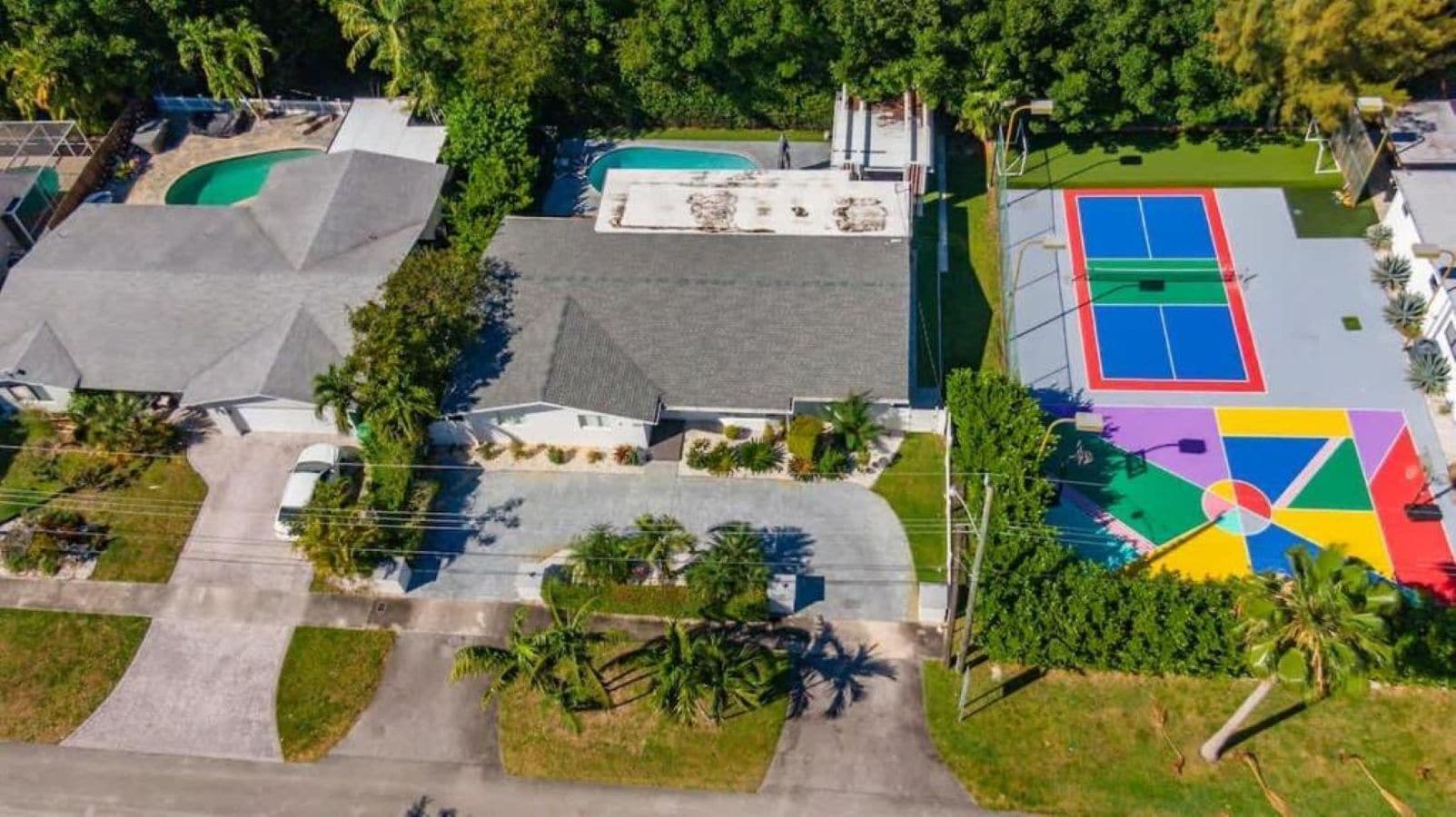 An aerial view of a residential area with a swimming pool, tennis court, and a house with a sloped roof.