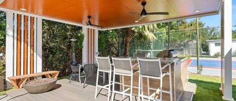 An outdoor patio with a wooden deck, white metal chairs, a round table, and a ceiling fan.