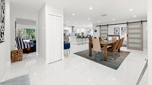 A modern kitchen with white cabinets, a dining area with a wooden table and chairs, and a sliding glass door leading to a patio. - Casa Oasis by Glam Homes (Ojus)