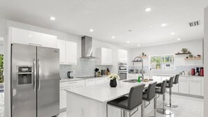 A modern kitchen with a stainless steel refrigerator, a central island with bar stools, and white cabinetry. - Casa Oasis by Glam Homes (Ojus)