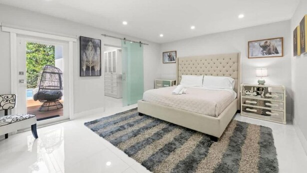 A bedroom with a bed, a chair, a nightstand, and a door leading to a balcony. - Casa Oasis by Glam Homes (Ojus)