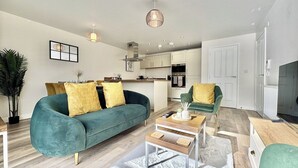 Una sala moderna con sofá, mesa de centro y sillón, una cocina al fondo y una puerta que da a un baño.
