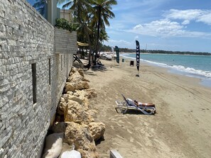 Una playa con un muro de piedra, palmeras y una persona recostada en una tumbona.