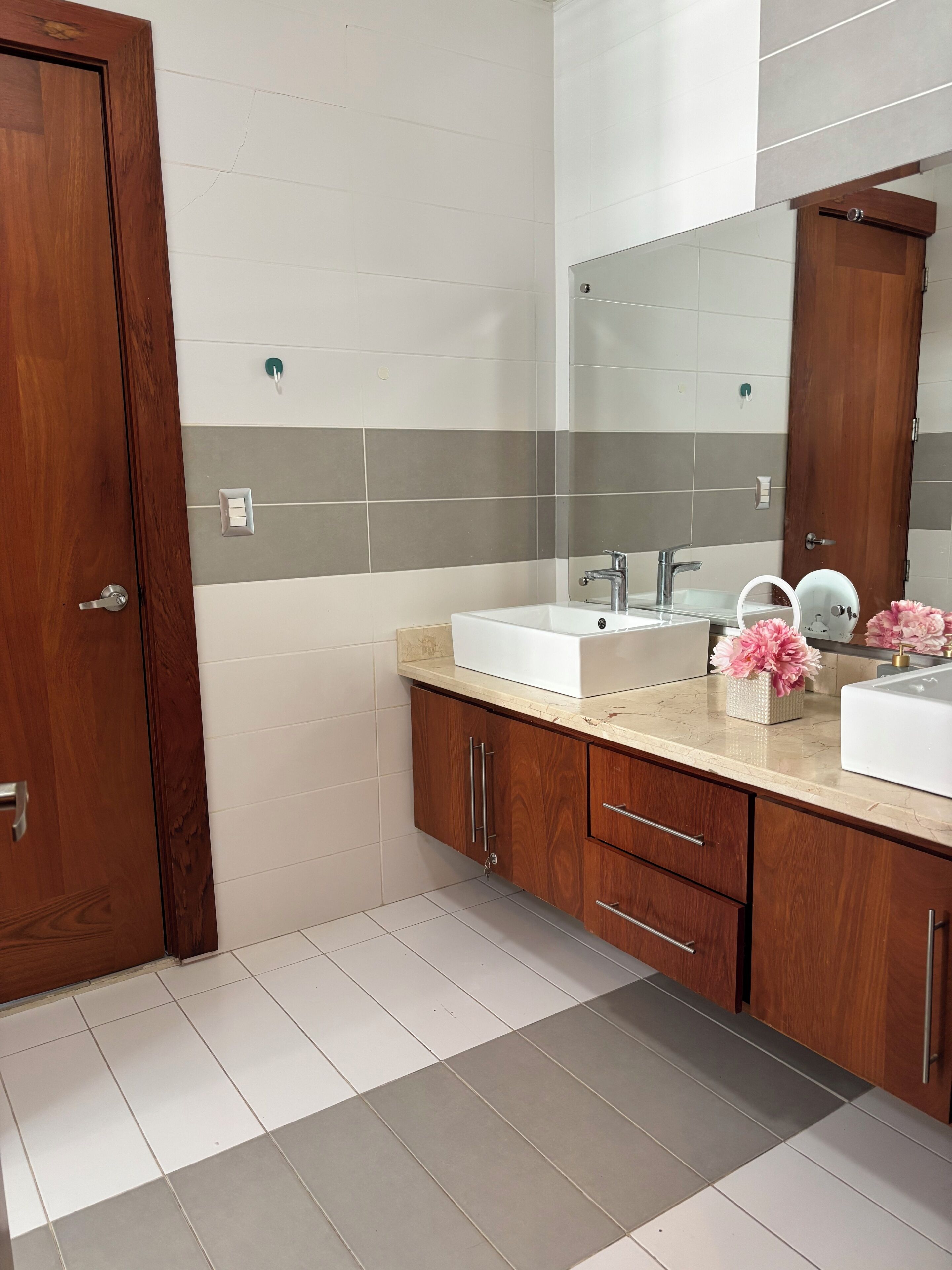 A bathroom with a wooden door, white and gray tiles, a white sink, a mirror, and a small vase with pink flowers.