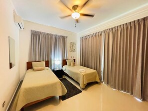 Habitación de hotel con dos camas, ventilador de techo, equipo de aire acondicionado y mesita de noche con lámpara.
