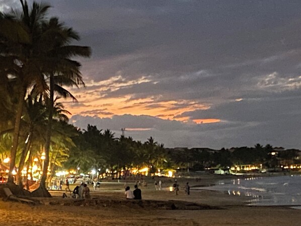 Escena de playa con palmeras, gente y un atardecer.