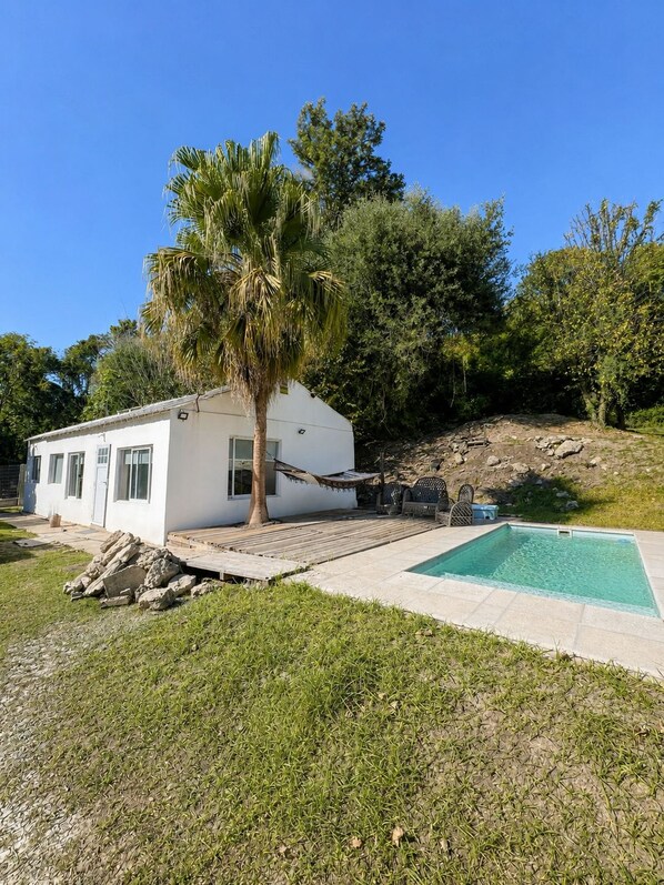 A white house with a pool, surrounded by greenery and a palm tree.