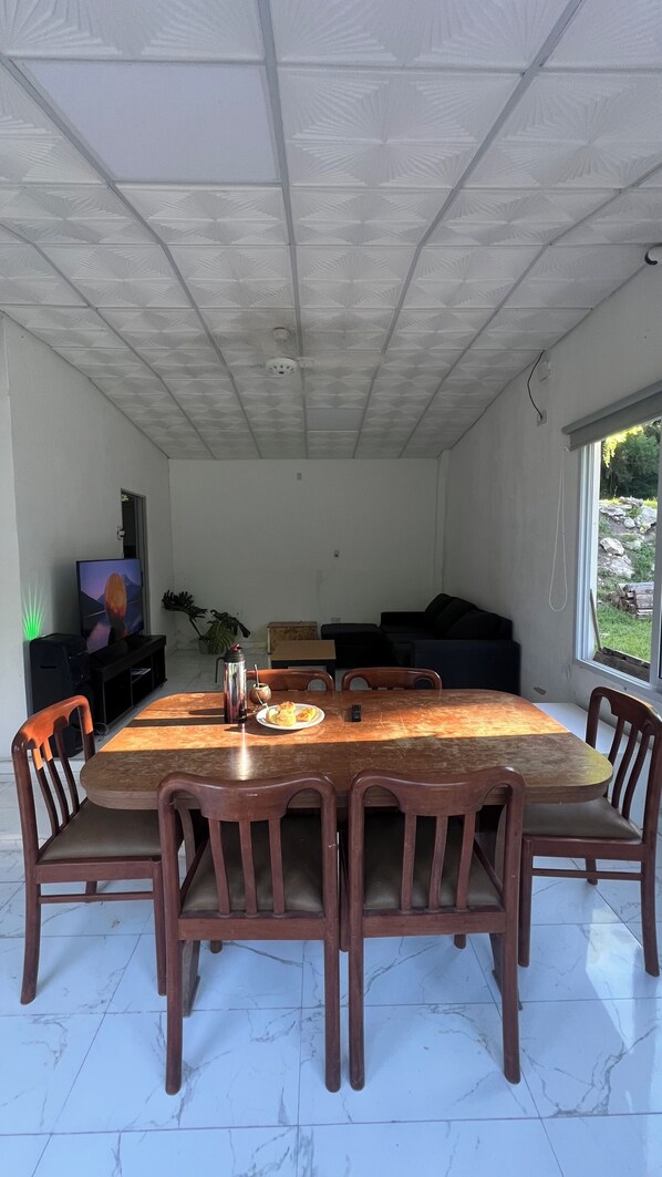 A dining area with a large wooden table, six chairs, a television, and a window with a view of greenery.