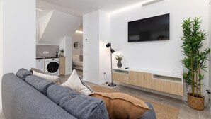 Una sala de estar moderna con un sofá gris, un televisor de pantalla plana, una planta en maceta y una cocina al fondo.