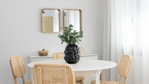 Un comedor con una mesa redonda blanca, cuatro sillas de mimbre, una planta en maceta en el centro, una lámpara colgante de fibras naturales y dos espejos adosados a la pared.