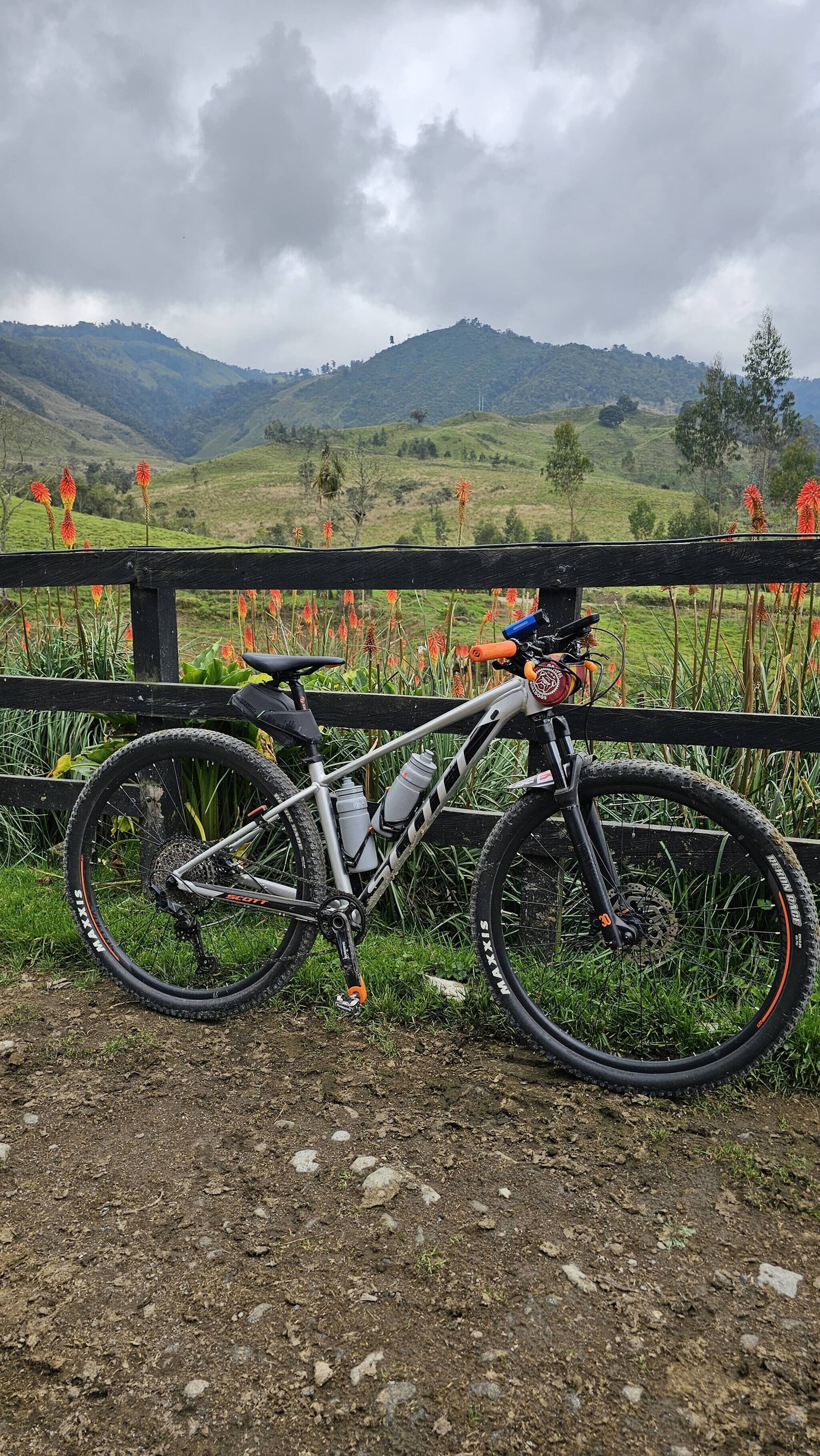 Una mountain bike parcheggiata su un sentiero sterrato con una recinzione di legno e vista su dolci colline verdi sullo sfondo.