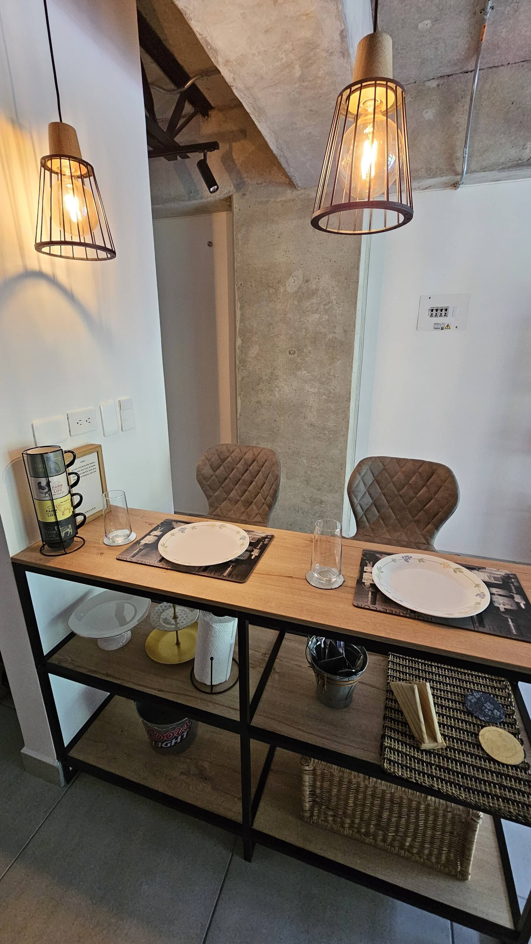 A dining area with a wooden table, two chairs, hanging pendant lights, and a wall-mounted electrical outlet.