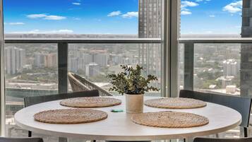 Une table ronde avec des sets de table en fibres tressées, une plante en pot et un porte-clés vert sur un balcon offrant une vue sur la ville.