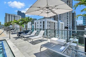 A rooftop lounge with sun loungers, a pool, and a glass balustrade overlooking a cityscape. - Designer Studio | 24th Floor Gym | Brickell Core (Miami)