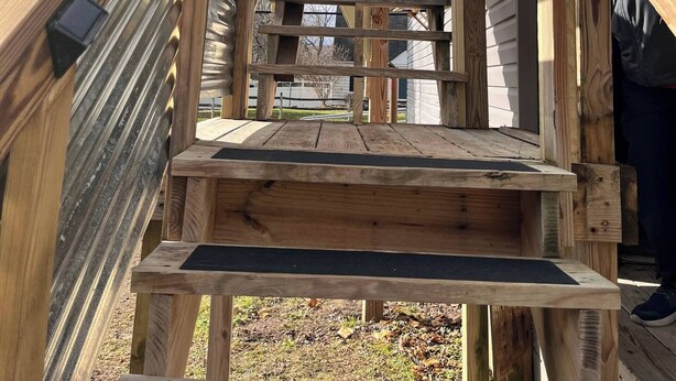 Eine Holztreppe führt zu einer weißen Tür mit einem kleinen Fenster, umgeben von einer Holzdielen-Terrasse.