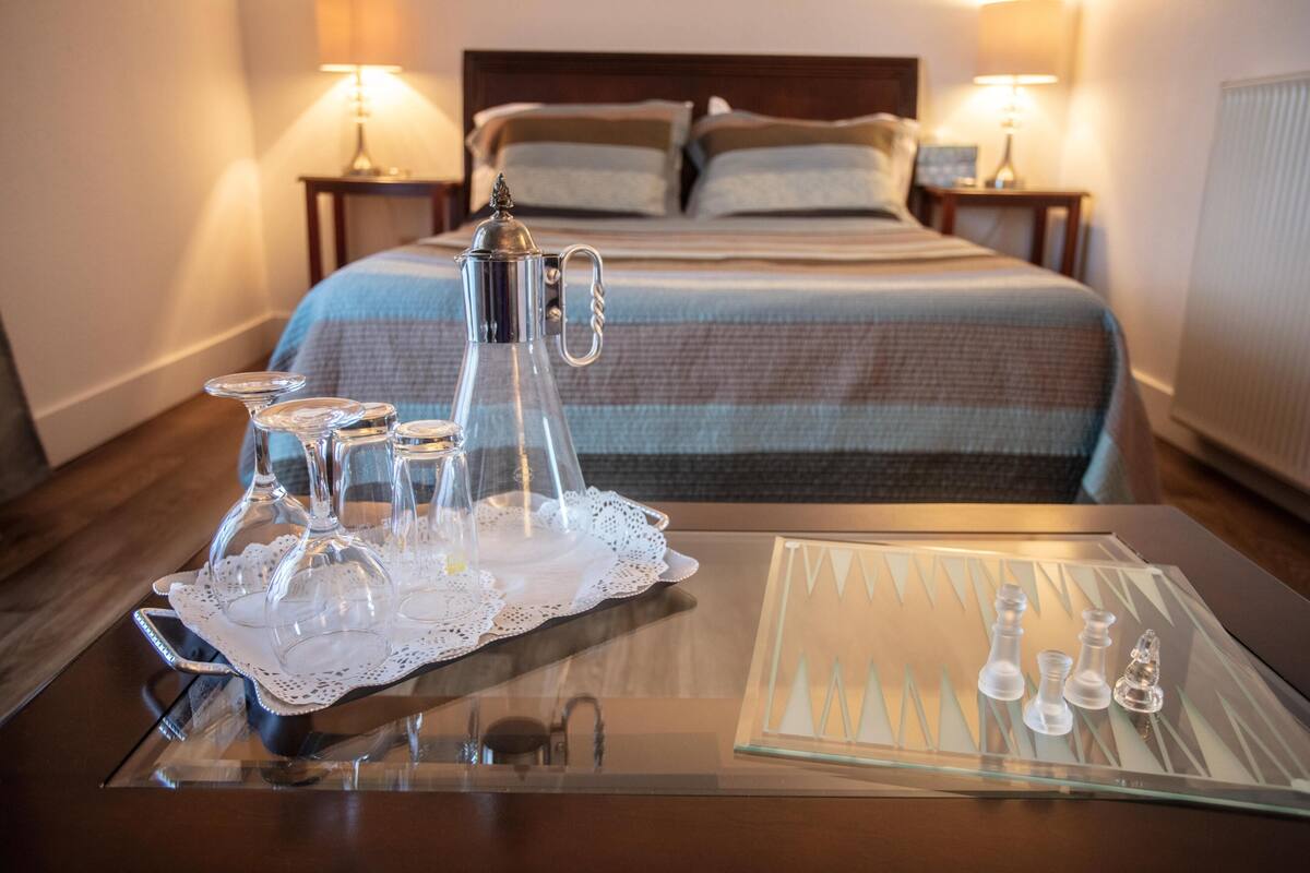 A neatly arranged table with a coffee pot, glasses, and a chessboard in a hotel room with a bed and nightstands in the background.