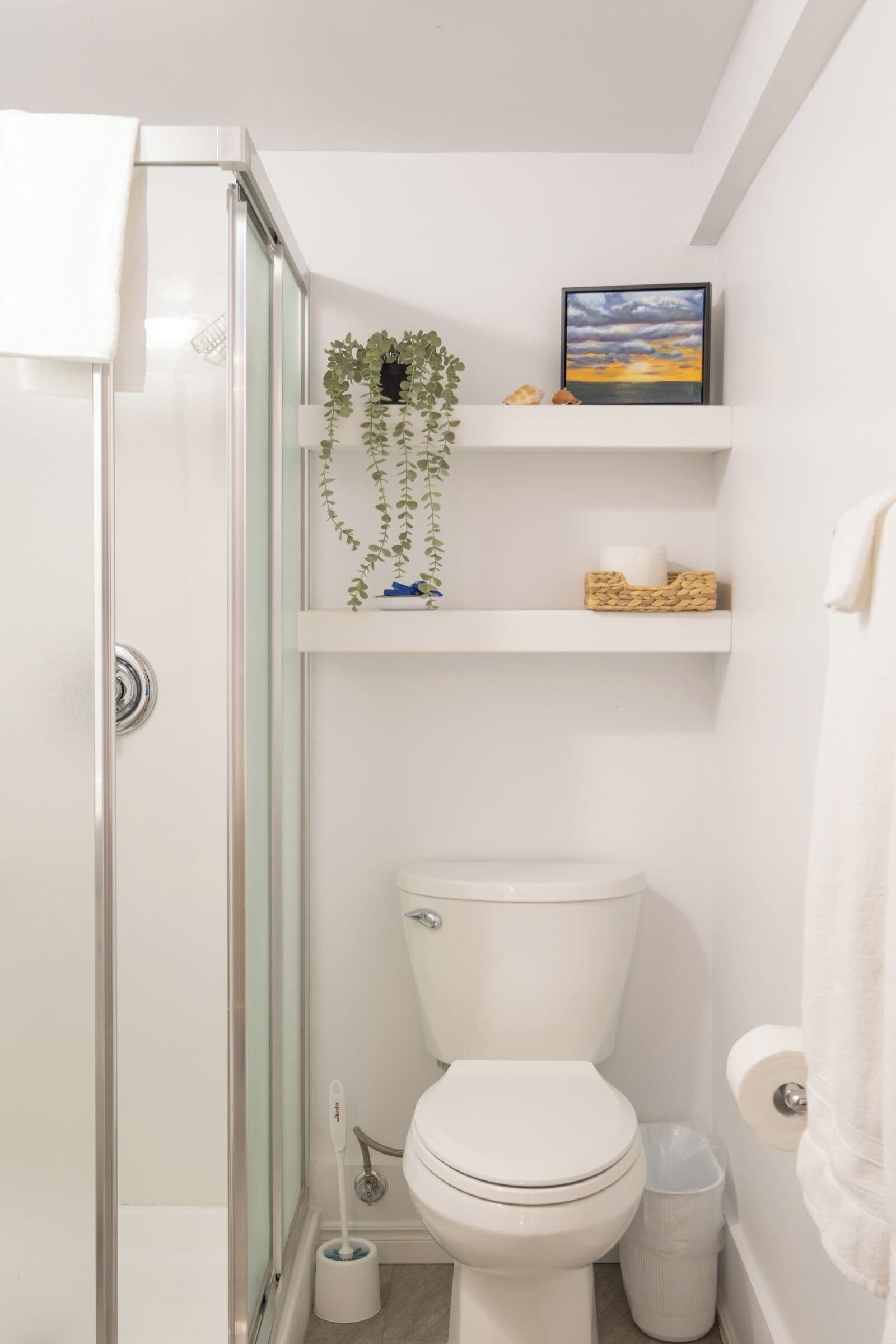 A bathroom with a glass shower enclosure, a toilet, a bidet, and a shelf with a plant and toiletries.
