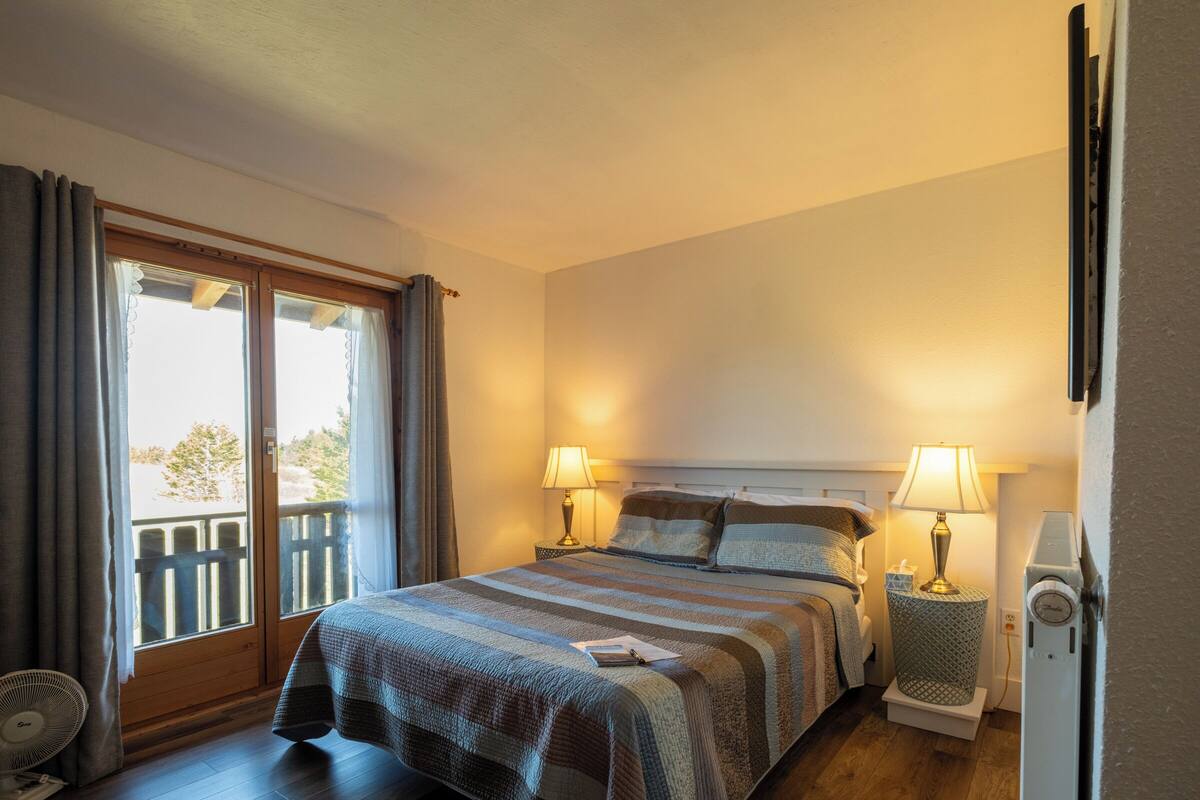 A hotel room with a bed, two bedside lamps, a heater, a fan, and a window with curtains.