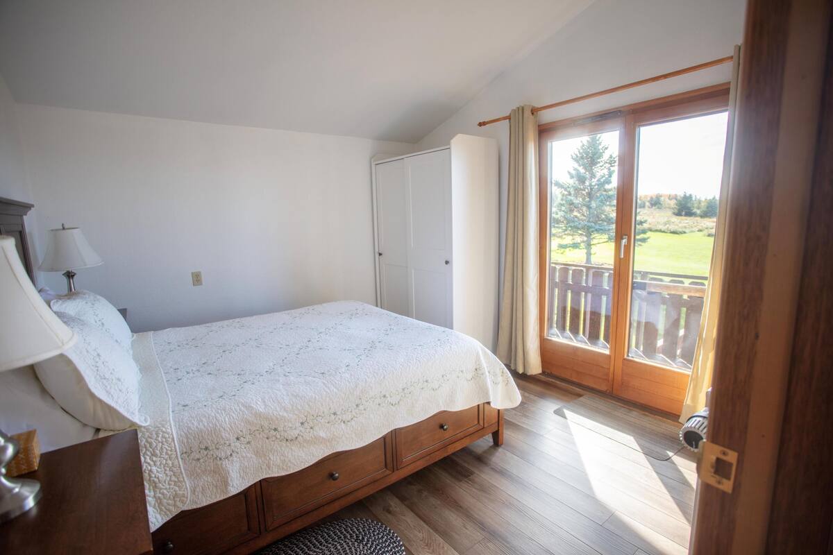 A bedroom with a bed, a nightstand, a lamp, a wooden door, and a view of a deck and trees.