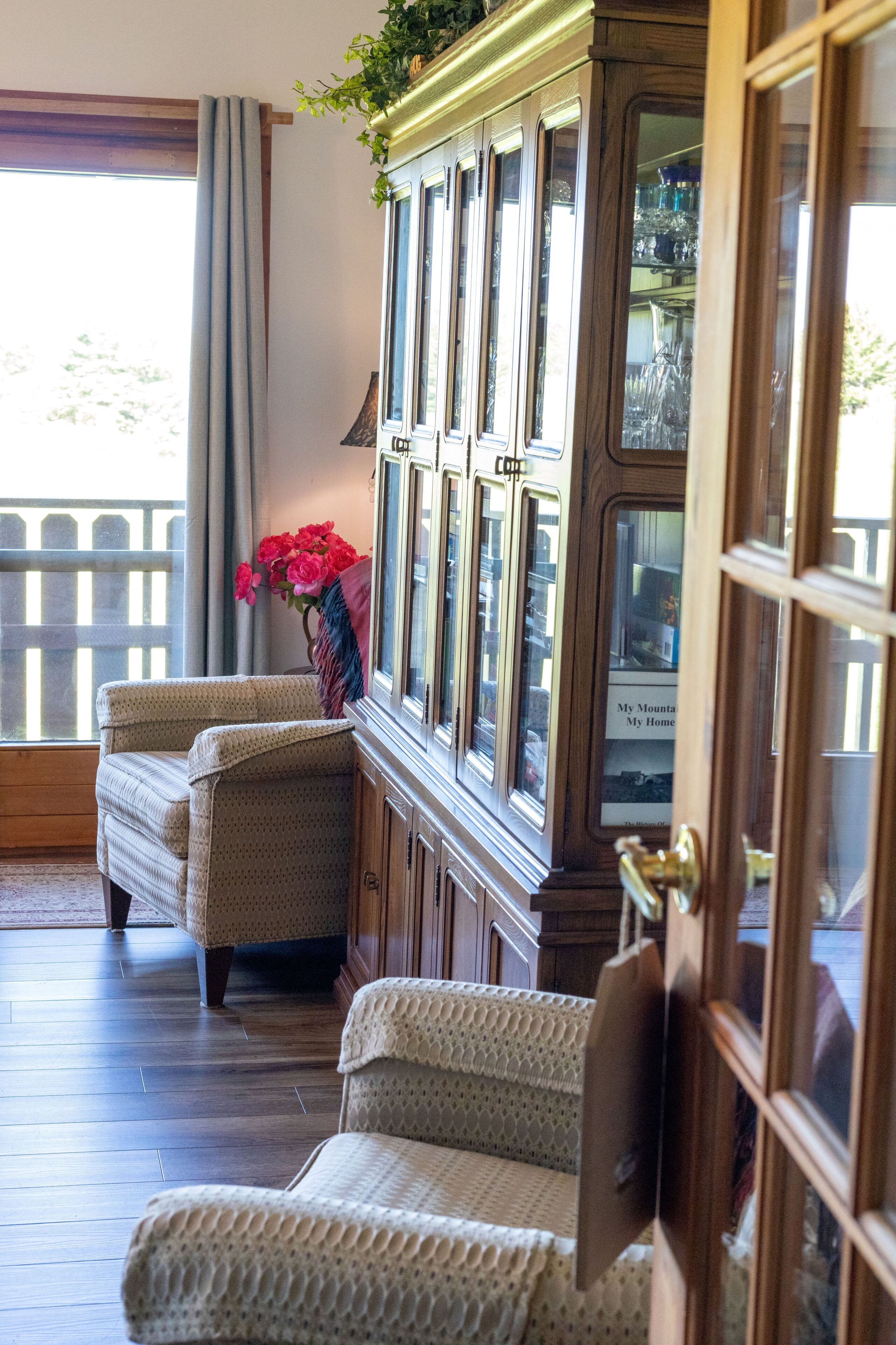 A room with a wooden cabinet, two patterned chairs, a window with curtains, and a view of a wooden deck.