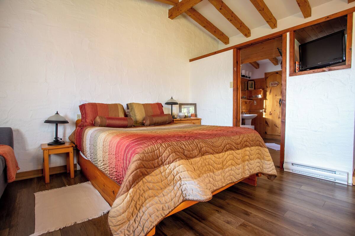 A bedroom with a wooden bed, a nightstand, a lamp, a window, and a television mounted on the wall.