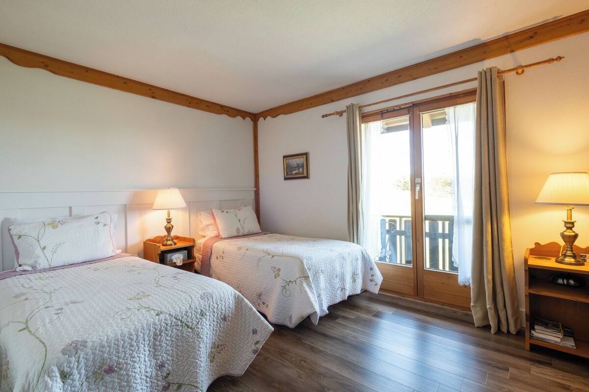 A room with two beds, wooden floors, a balcony with curtains, and a small table with a lamp.