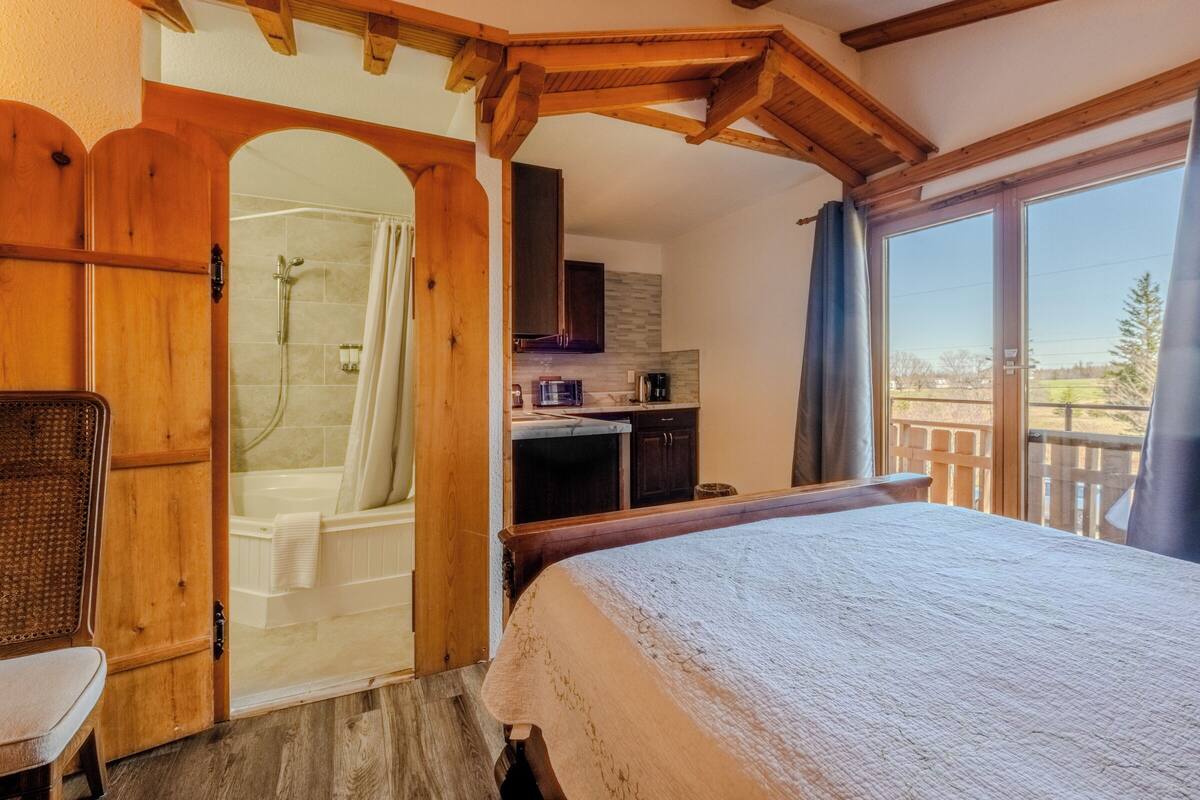 A bedroom with a large bed, wooden ceiling beams, a shower room, and a view of the outdoors.