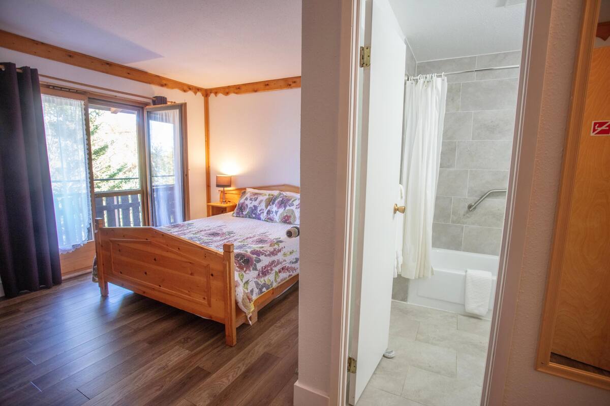A bedroom with a wooden bed, a nightstand, a lamp, and a view of the outside.