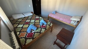 Une chambre avec deux lits : l’un est recouvert d’une couvre-lit à motifs et agrémenté d’oreillers, l’autre est recouvert d’une simple couverture rose. On y trouve une petite table en bois et un radiateur posé sur le sol.