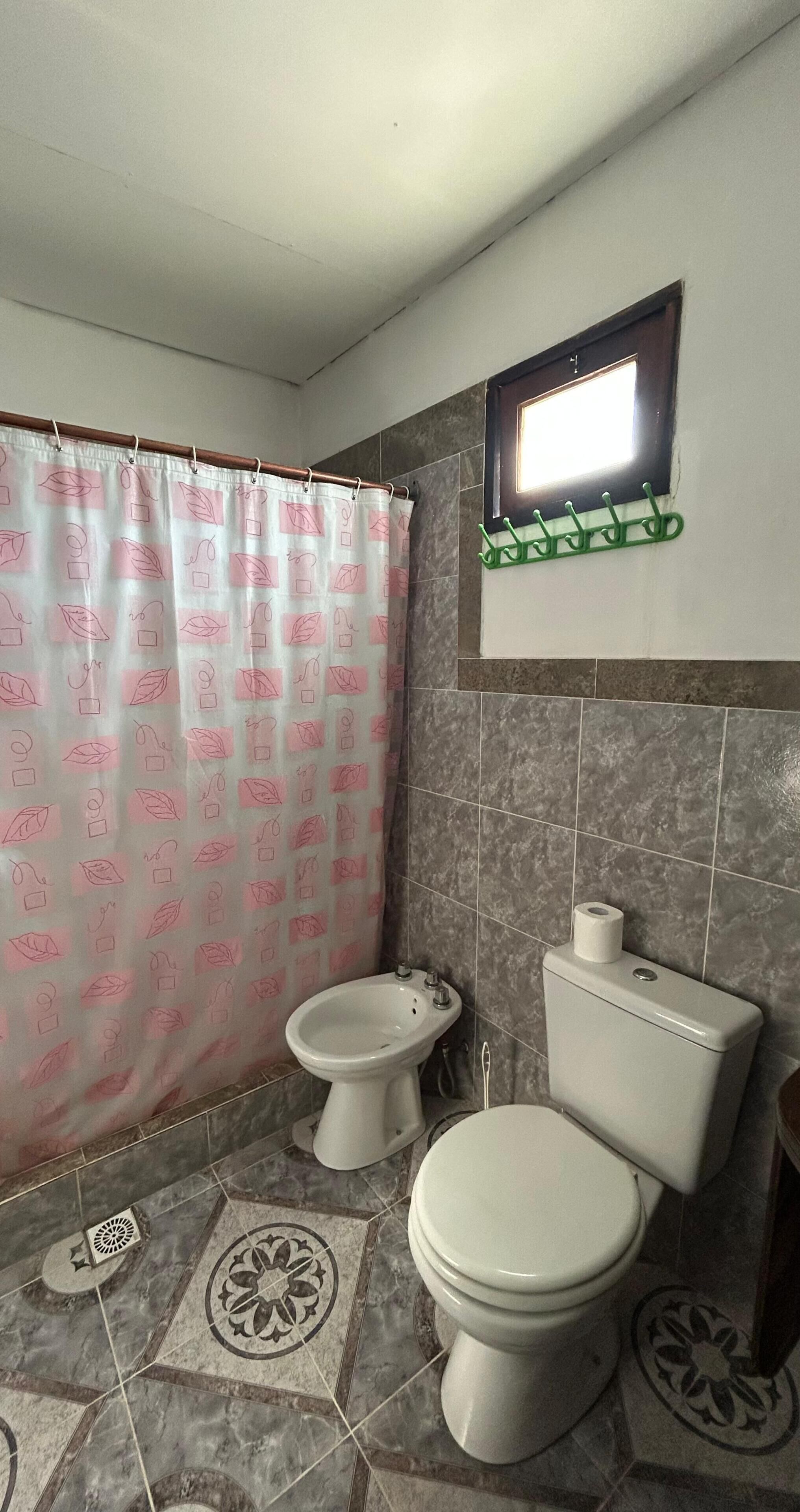 A bathroom with two toilets, a shower curtain with a pattern, a green wall-mounted rack, and patterned floor tiles.
