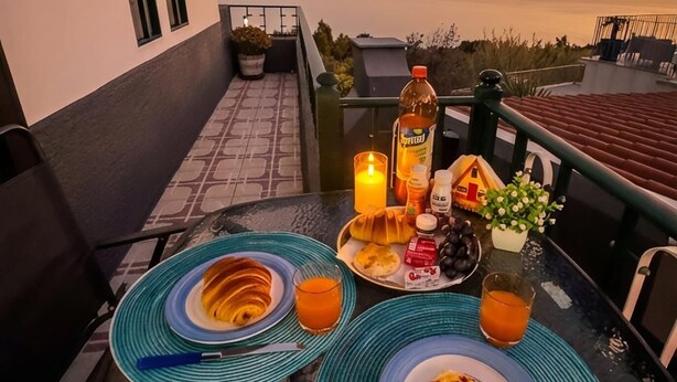 Een tafel gedekt met ontbijtartikelen, een kaars en een fles, op een balkon met uitzicht op een zonsondergang.
