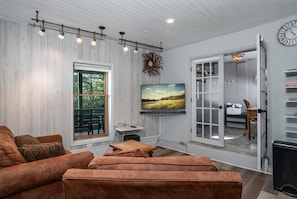 A living room with a large TV, a wooden coffee table, a brown sofa, and a door leading to a kitchen area. - 'Amongst the Clouds,' a one-of-a-kind retreat. (Blowing Rock)