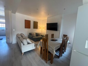 A living room with a sofa, a dining table with chairs, a flat-screen TV mounted on the wall, and a lamp.