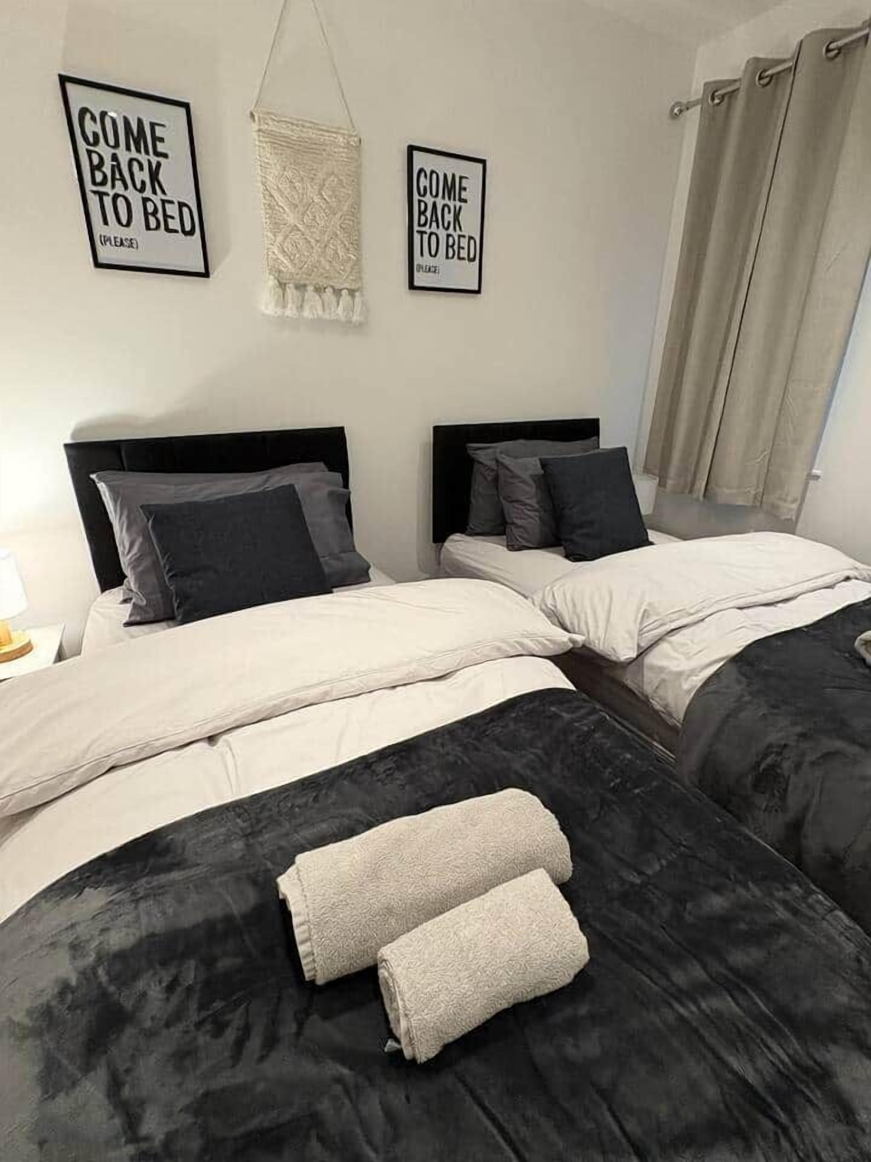 A hotel room with two beds, each with black headboards and white bedding. There are two signs on the wall, one above each bed, reading 'COME BACK TO BED'.