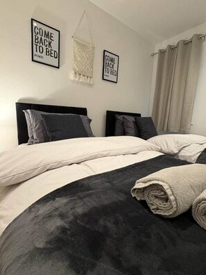 Een tweepersoonsbed met een zwart hoofdbord en een beige deken, twee kussens en een opgerolde handdoek. Twee borden aan de muur met de tekst 'COME BACK TO BED (PLEASE)'.
