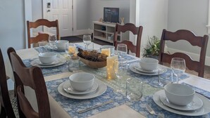 Un coin repas avec une table dressée pour quatre personnes, des chaises, un vase décoratif, des bougies et une fenêtre avec des stores.