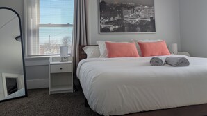 Une chambre à coucher bien rangée, avec un lit, des oreillers, une table de chevet, un miroir et une fenêtre avec des stores.