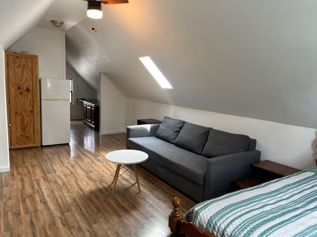 Ein Schlafzimmer im Loft-Stil mit einem Bett, einem grauen Sofa, einem kleinen runden Tisch, einer Holztür und einer Küchenzeile im Hintergrund.