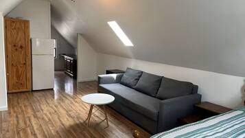 Ein Schlafzimmer im Loft-Stil mit einem Bett, einem grauen Sofa, einem kleinen runden Tisch, einer Holztür und einer Küchenzeile im Hintergrund.