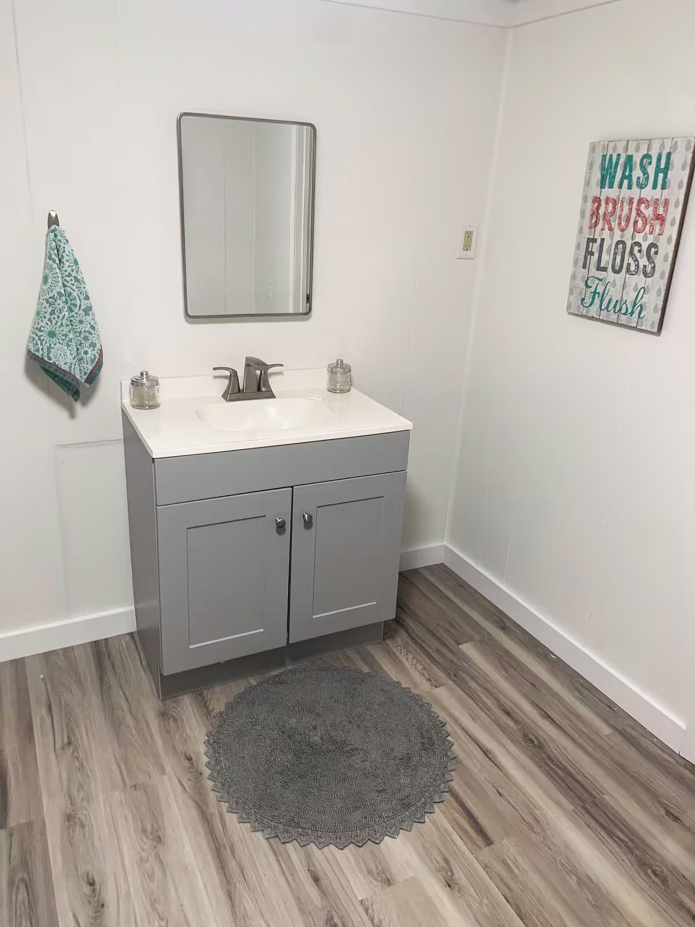 Une salle de bain avec un lavabo blanc, des meubles gris, un miroir, une serviette et un tapis.