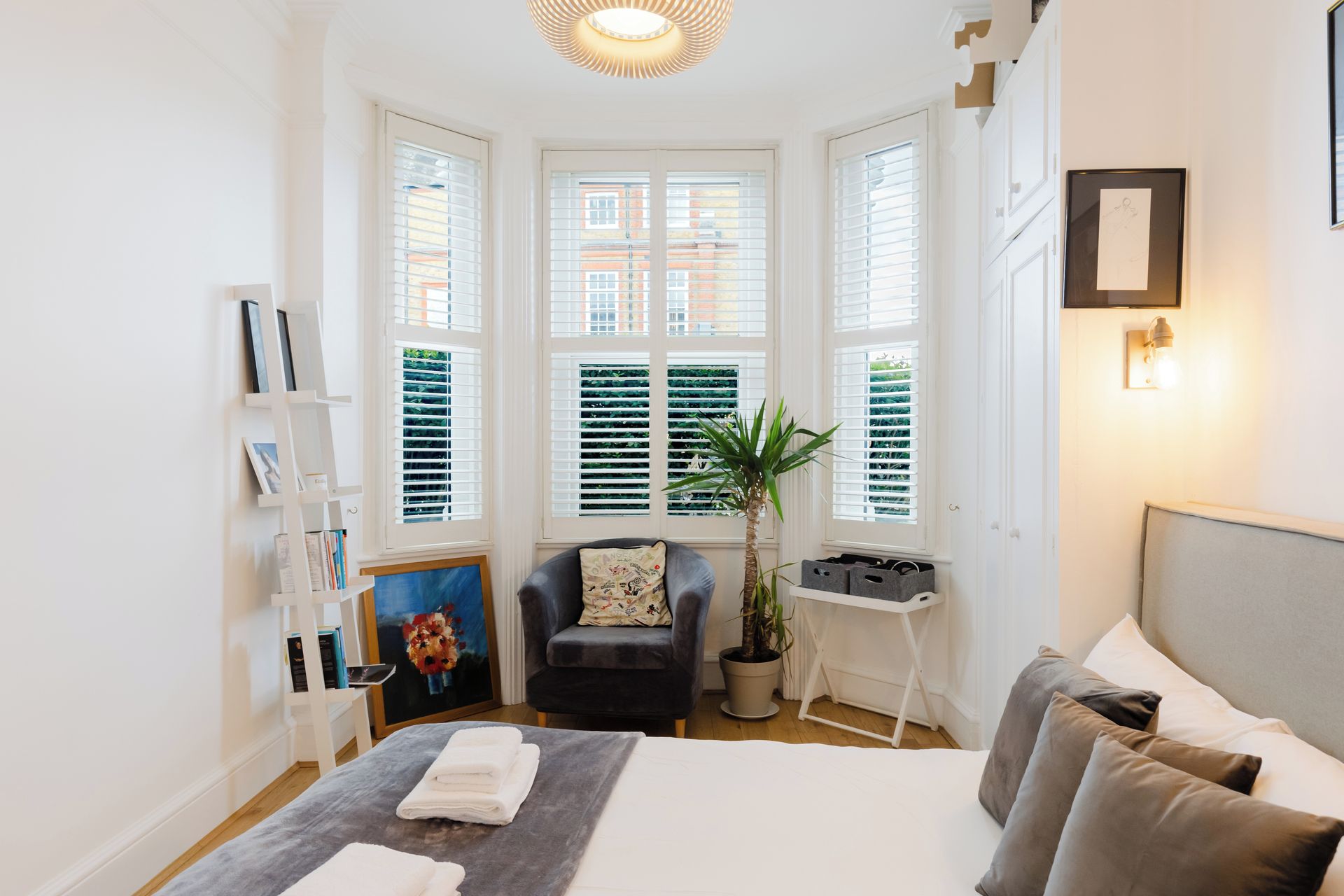 A bedroom with a bed, a chair, a desk, a potted plant, and a window with blinds.
